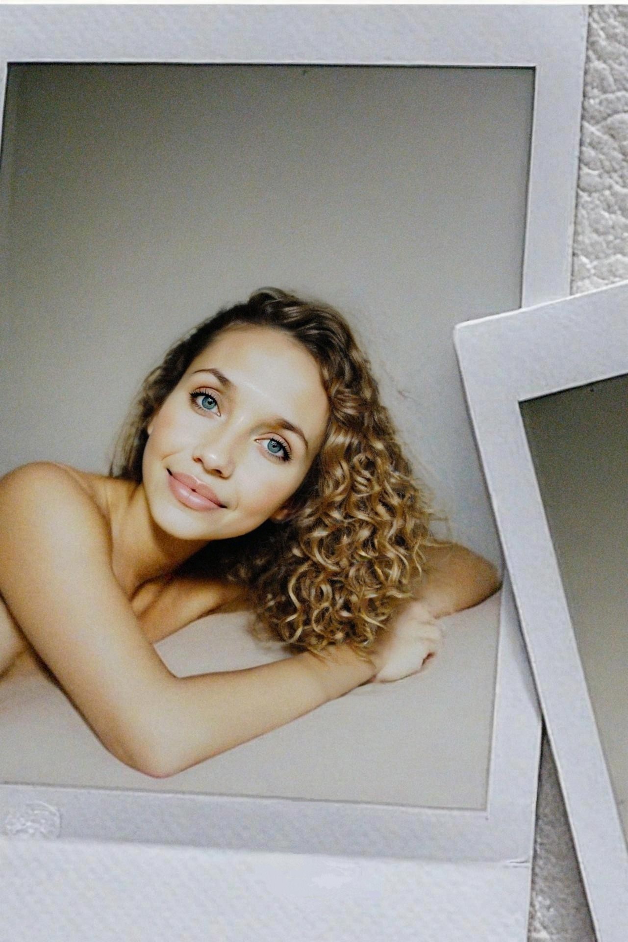 A close-up portrait of a woman with curly hair and blue eyes, smiling warmly while resting on her arms against a neutral background.